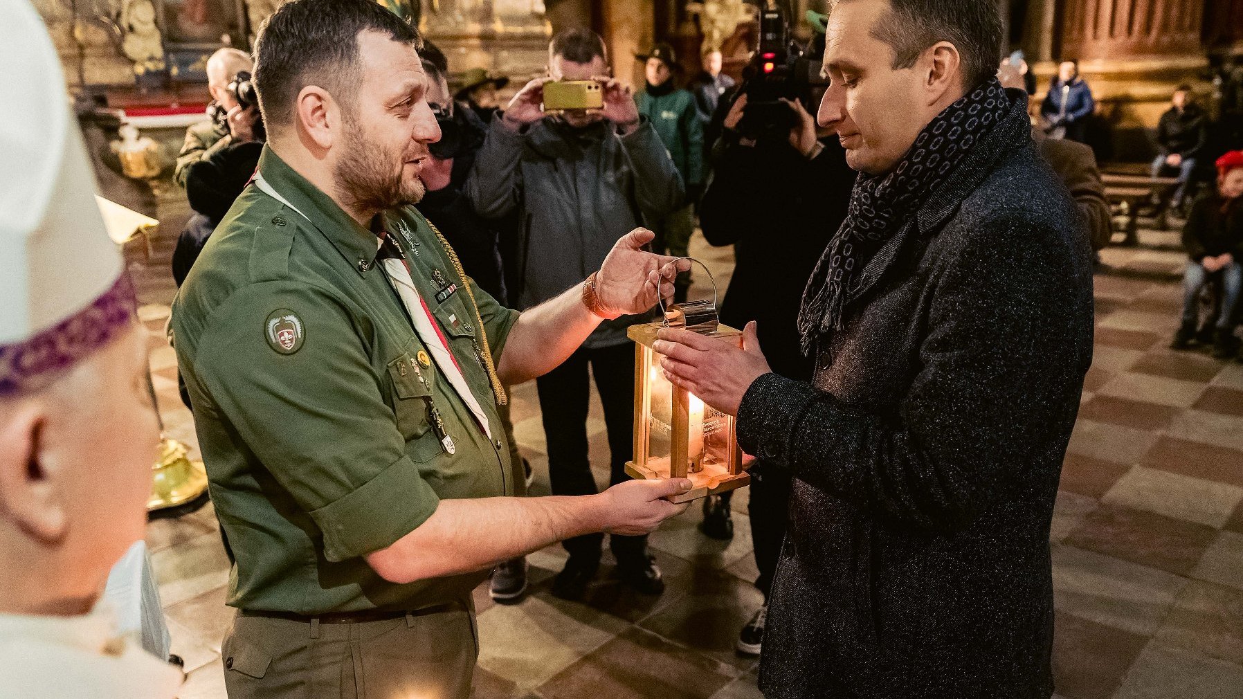 Osoba w zielonym mundurze, najprawdopodobniej harcerz, przekazuje latarnię z zapaloną świecą drugiej osobie w ciemnym płaszczu i szaliku. Ceremonia odbywa się w kościele lub podobnym miejscu sakralnym, co sugerują ozdobne elementy architektoniczne i obecność artefaktów religijnych. W tle widoczne są inne osoby, niektóre z nich robią zdjęcia lub nagrywają wydarzenie.