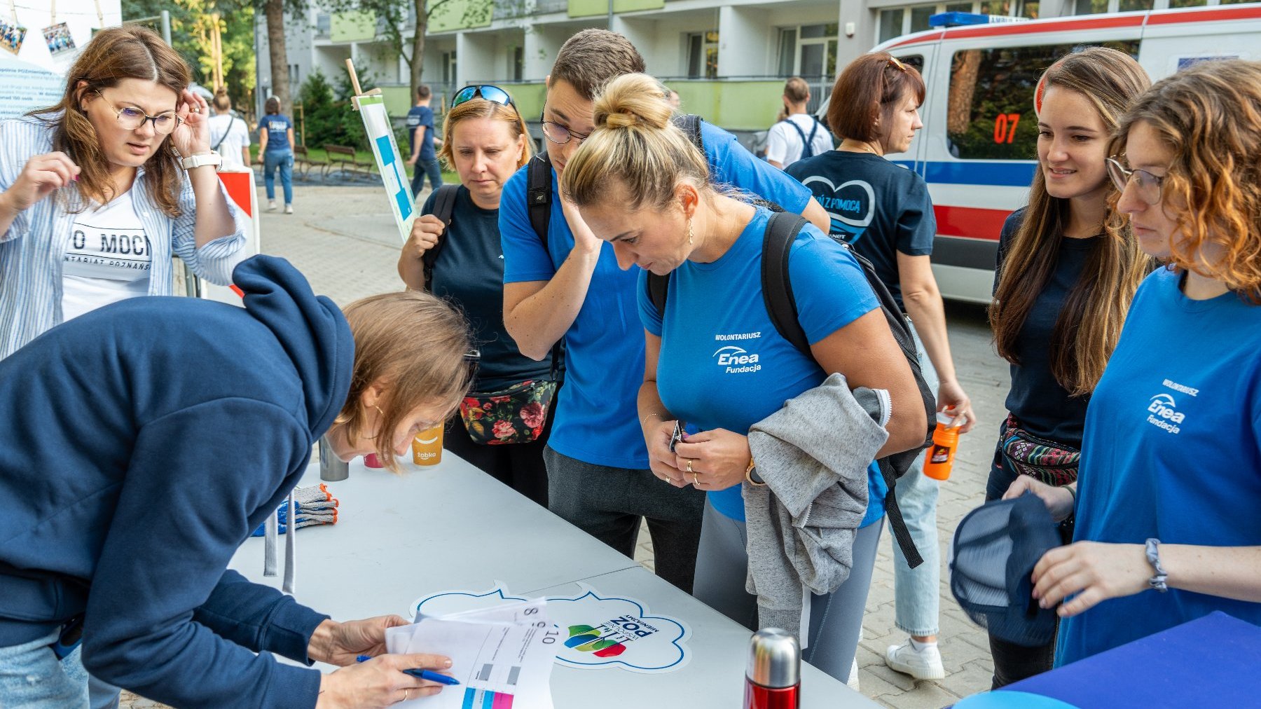 Na zdjęciu grupa wolontariuszy w niebieskich koszulkach, pochylają się nad stolikiem