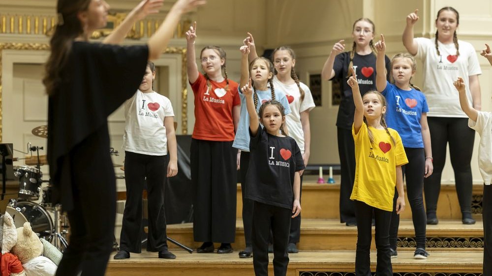 Na zdjęciu dzieci i młodzież na próbie chóru, ubrani w kolorowe t-shirty, podnoszą prawe ręce w górę. Naprzeciwko nich stoi dyrygentka.