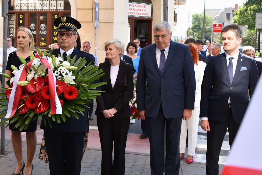 Na zdjęciu kilka osób składających wiązankę kwiatów, na pierwszym planie strażnik miejski z kwiatami - grafika artykułu