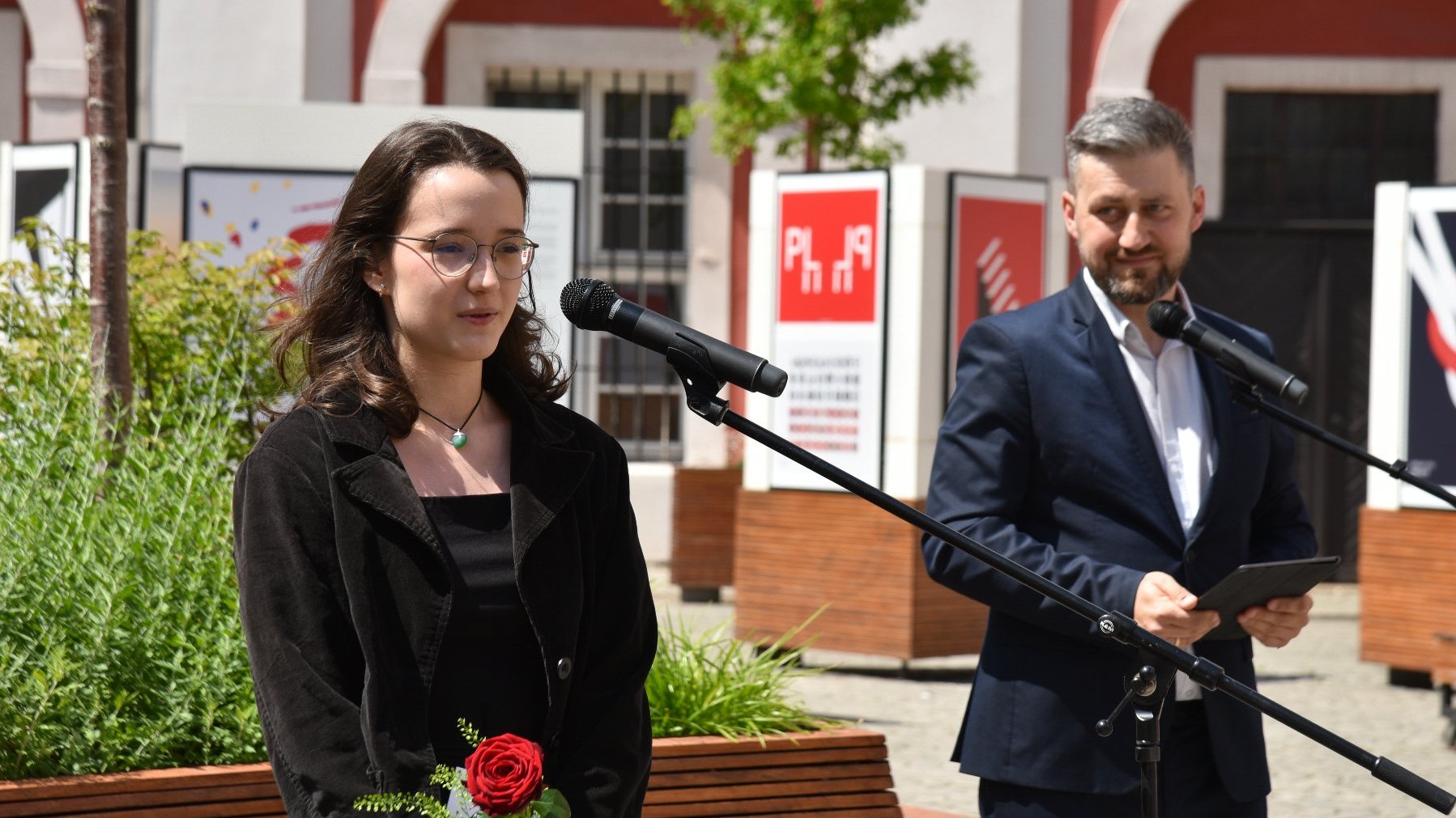 Na zdjęciu młoda kobieta trzymająca różę, przy mikrofonie, za nią mężczyzna