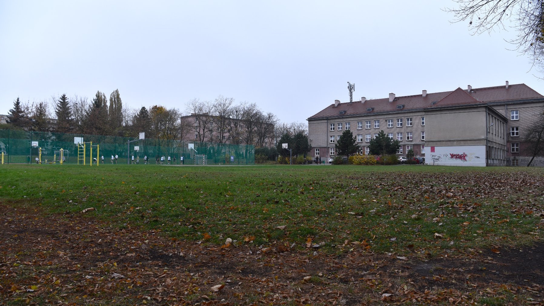 Zdjęcie przedstawia uczniów grających w piłkę na boisku oraz budynek szkoły.