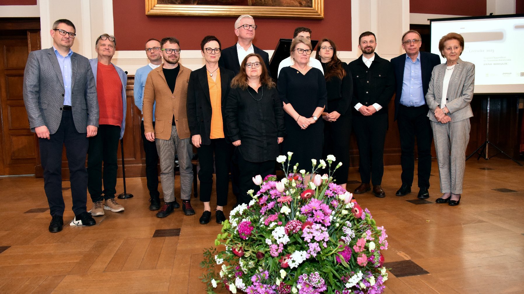 Zdjęcie przedstawia grupę osób stojących w Auli Lubrańskiego pod obrazem wiszącym na czerwonej ścianie - grafika artykułu