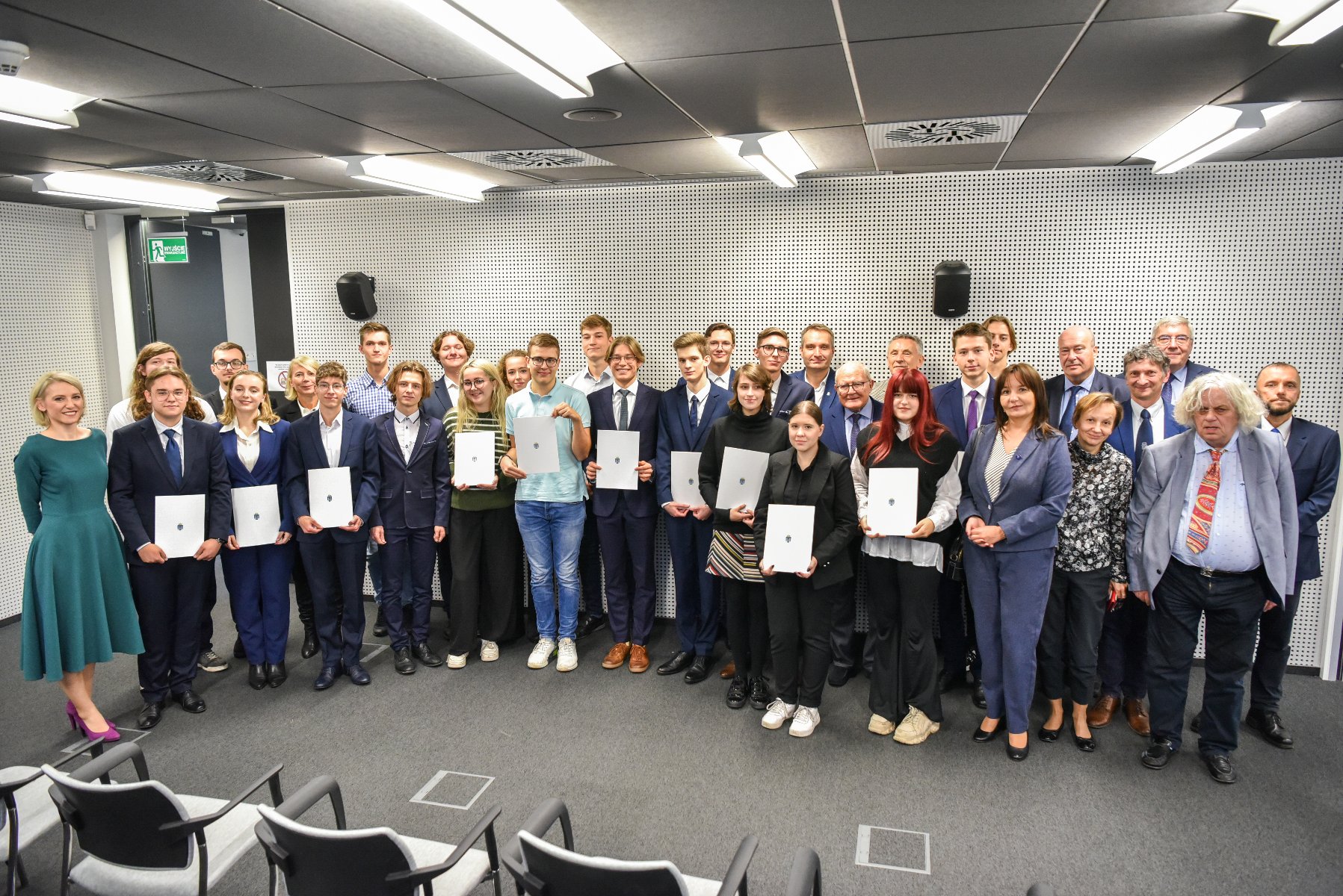 Zdjęcie przedstawia grupę elegancko ubranych osób stojących w sali konferencyjnej - grafika artykułu