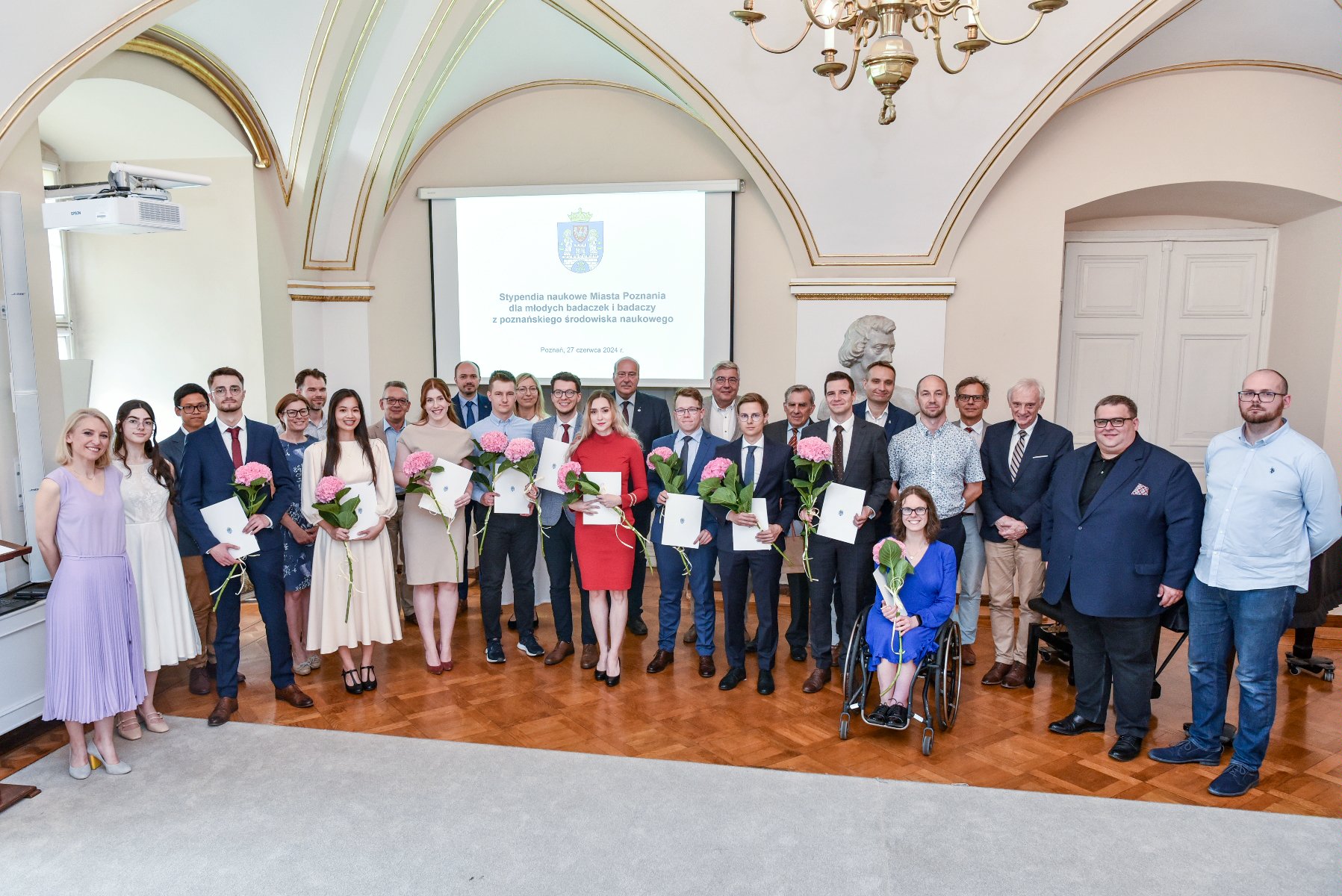 zdjęcie przedstawia grupę elegancko ubranych osób w sali urzędu miasta. W pierwszym rzędzie znajdują się laureatki i laureaci stypendium, którzy trzymają w rękach dyplomy i kwiaty. - grafika artykułu