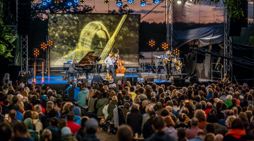 Na zdjęciu ludzie słuchający muzyki, w tle ustawiona w plenerze scena, na niej muzycy - grafika artykułu