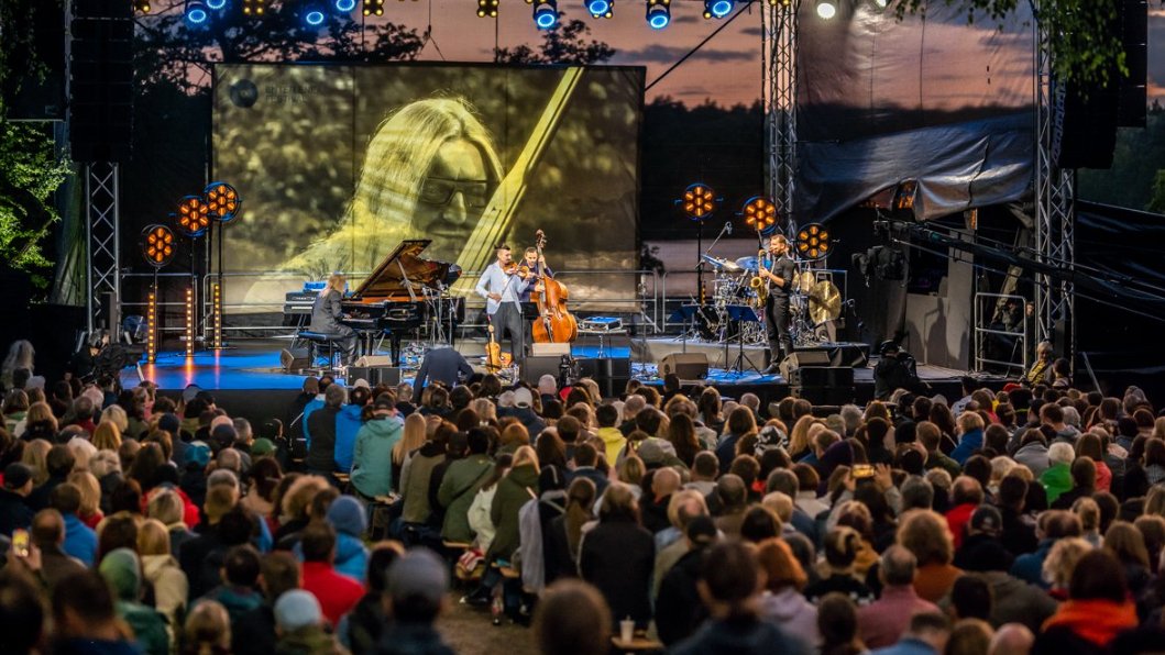 Na zdjęciu ludzie słuchający muzyki, w tle ustawiona w plenerze scena, na niej muzycy