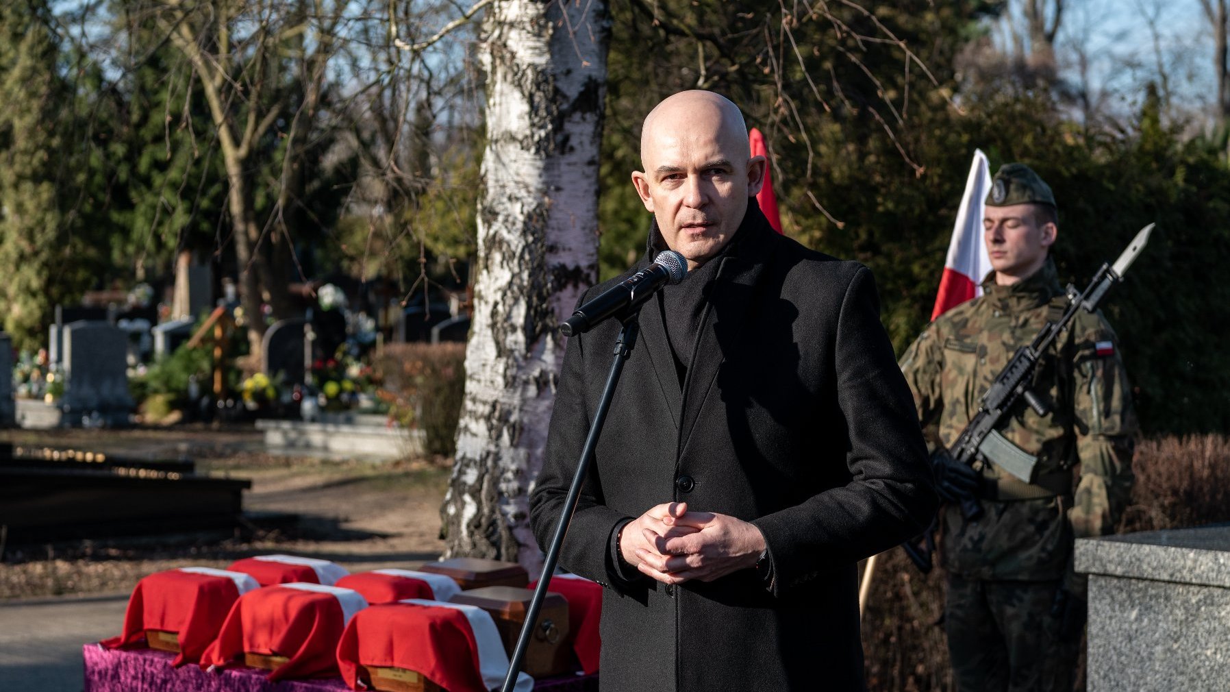 Na zdjęciu mężczyzna przy mikrofonie, za nim urny i żołnierz