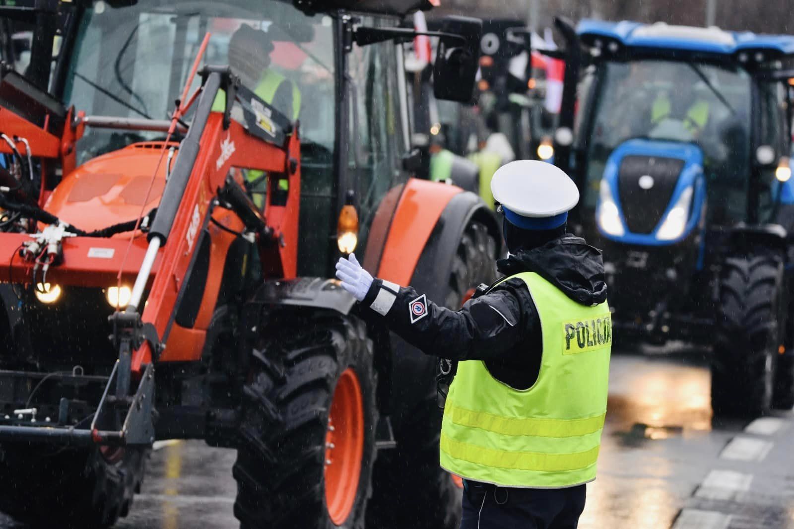 Zdjęcie przedstawia ciągniki i policjanta kierującego ruchem. - grafika artykułu