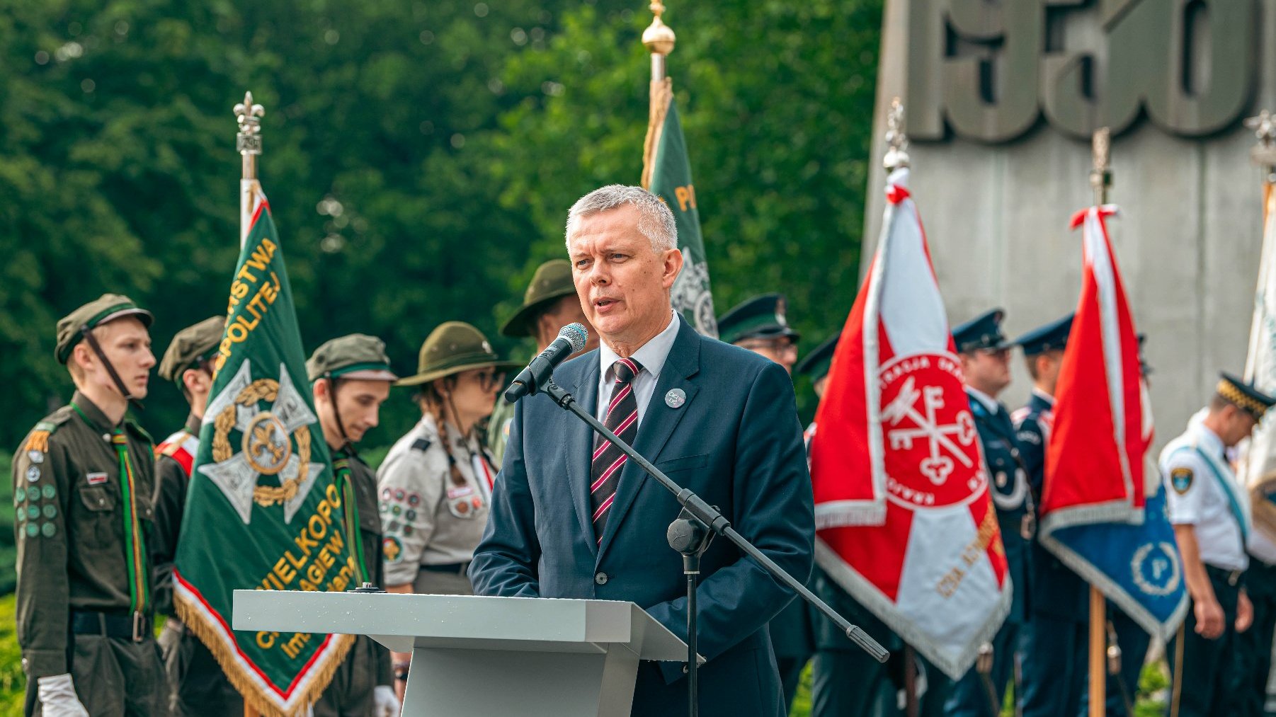 Na zdjeciu mężczyzna przy mównicy, w tle pomnik i poczty sztandarowe