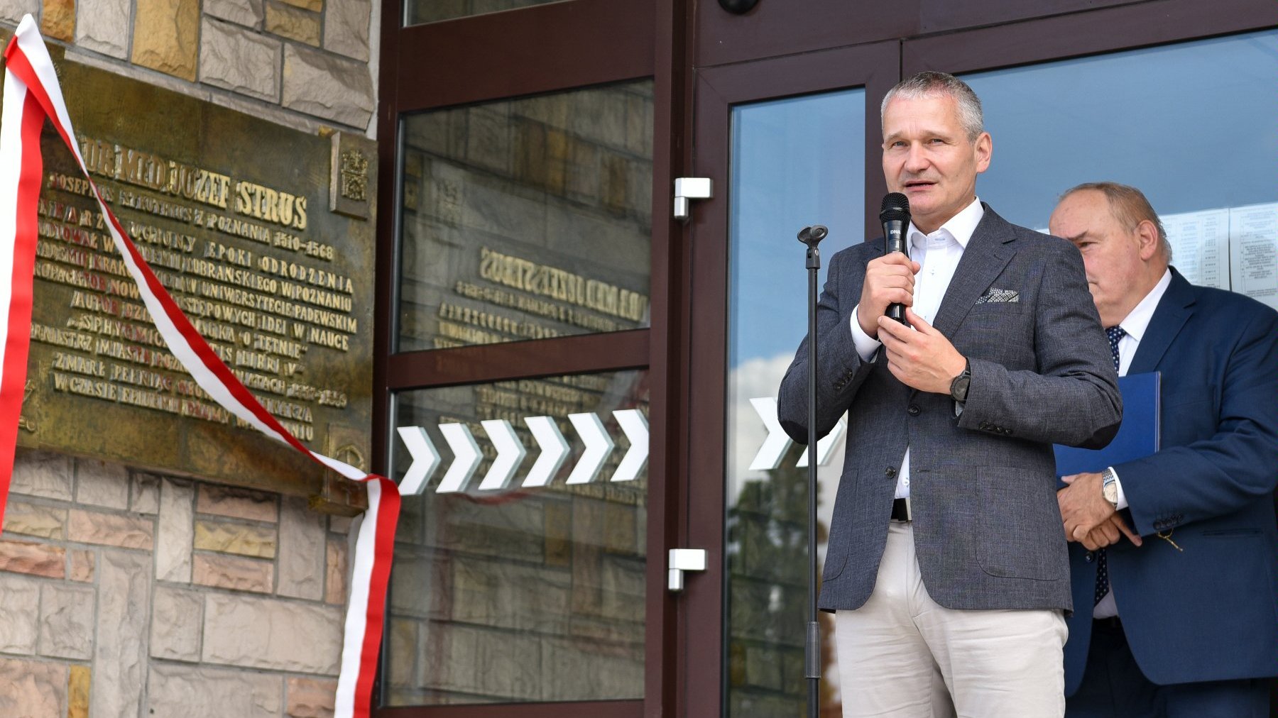 Na zdjeciu dwóch mężczyzn stojących obok tablicy, jeden z nich mówi coś do mikrofonu