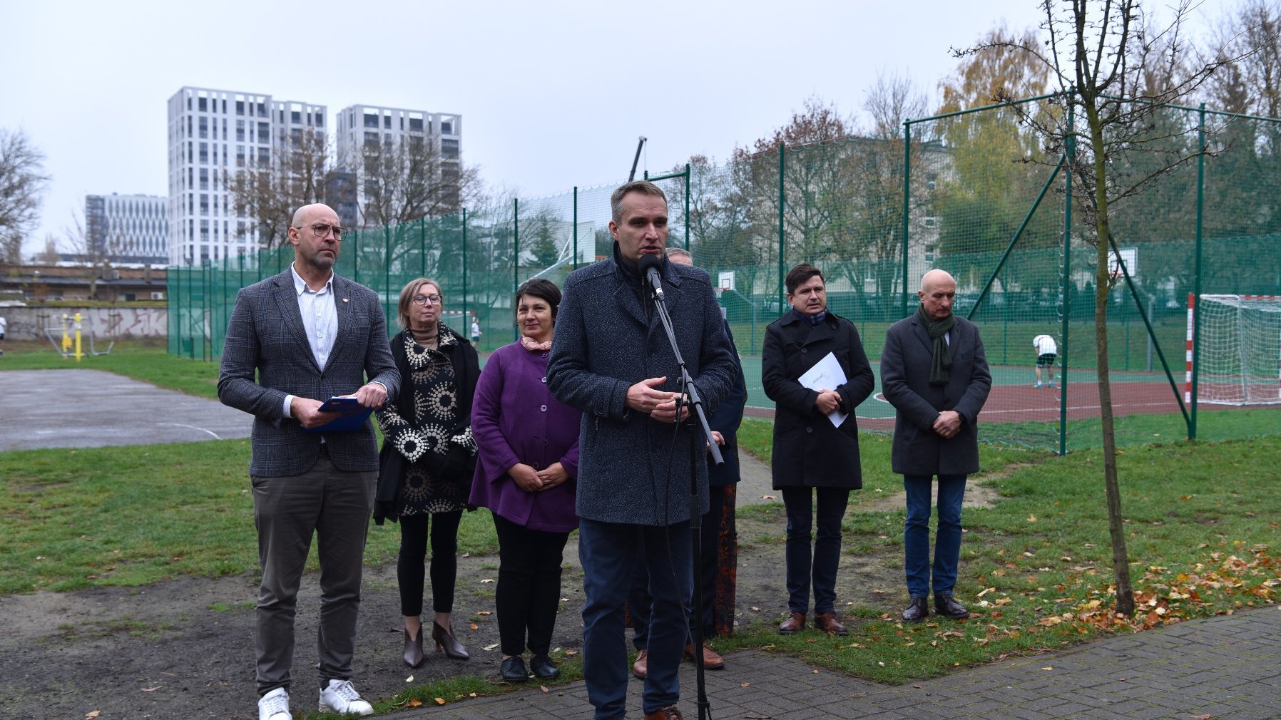 Zdjęcie przedstawia uczestników konferencji prasowej. Do mikrofonu mówi mężczyzna. Za ich plecami znajduje się boisko.