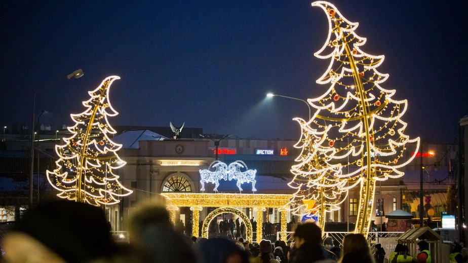 Widok na oświetlony iluminacjami teren targów. Po bokach widać iluminacje w kształcie choinek, w tle budynek targów i trykające się koziołki.