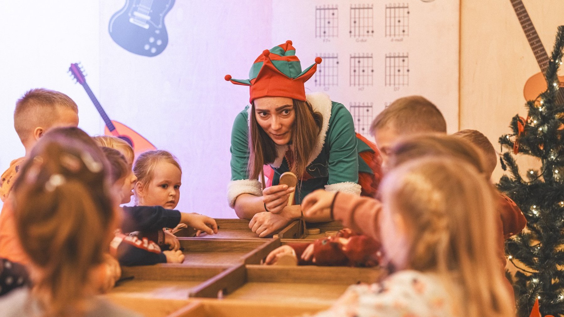 Kobieta w stroju elfa nachyla się nad stołem z żetonem w ręce. Dookoła niego stoją dzieci uczestniczące w grze.