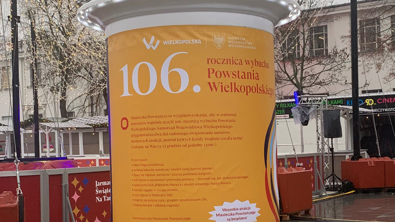 Na słupie ogłoszeniowym widać duży plakat z informacjami o 106. rocznicy wybuchu Powstania Wielkopolskiego i utworzeniu Miasteczka Powstańczego.