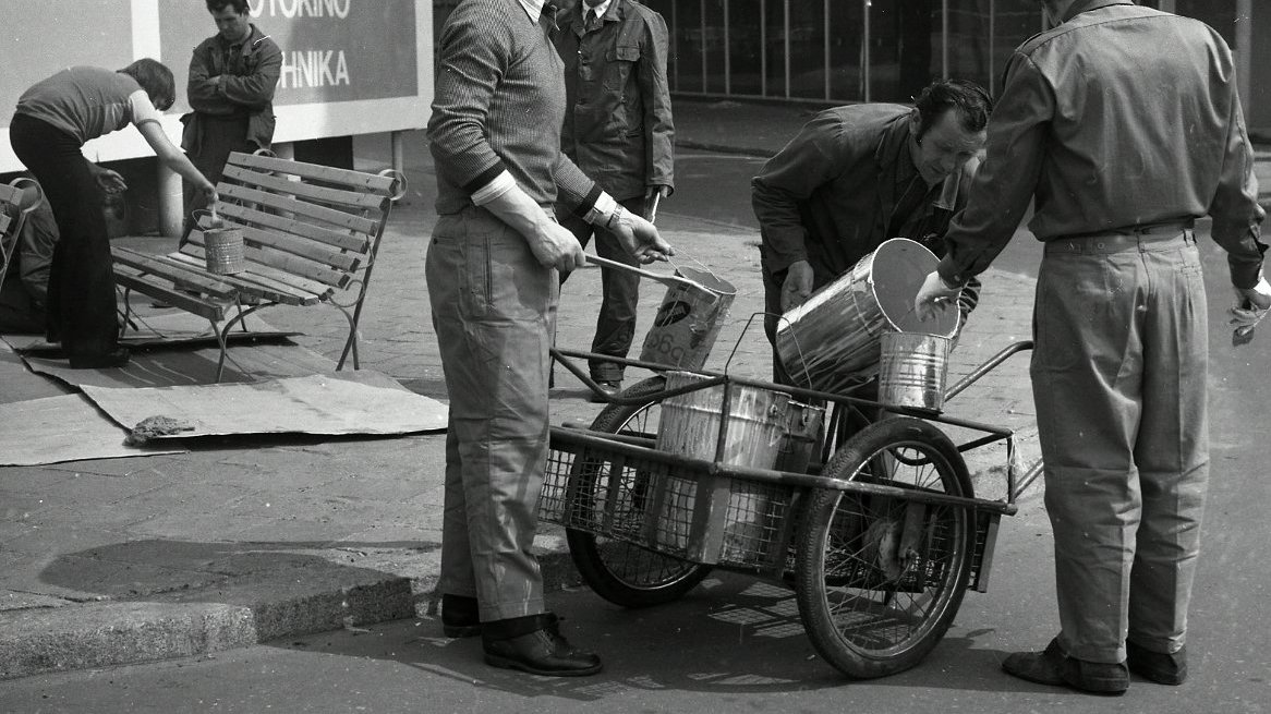 Black and white picture of a few men who are renovating a bench - one of them is paining it, one is standing by a bench, three other men are pouring the paint from big to small can.