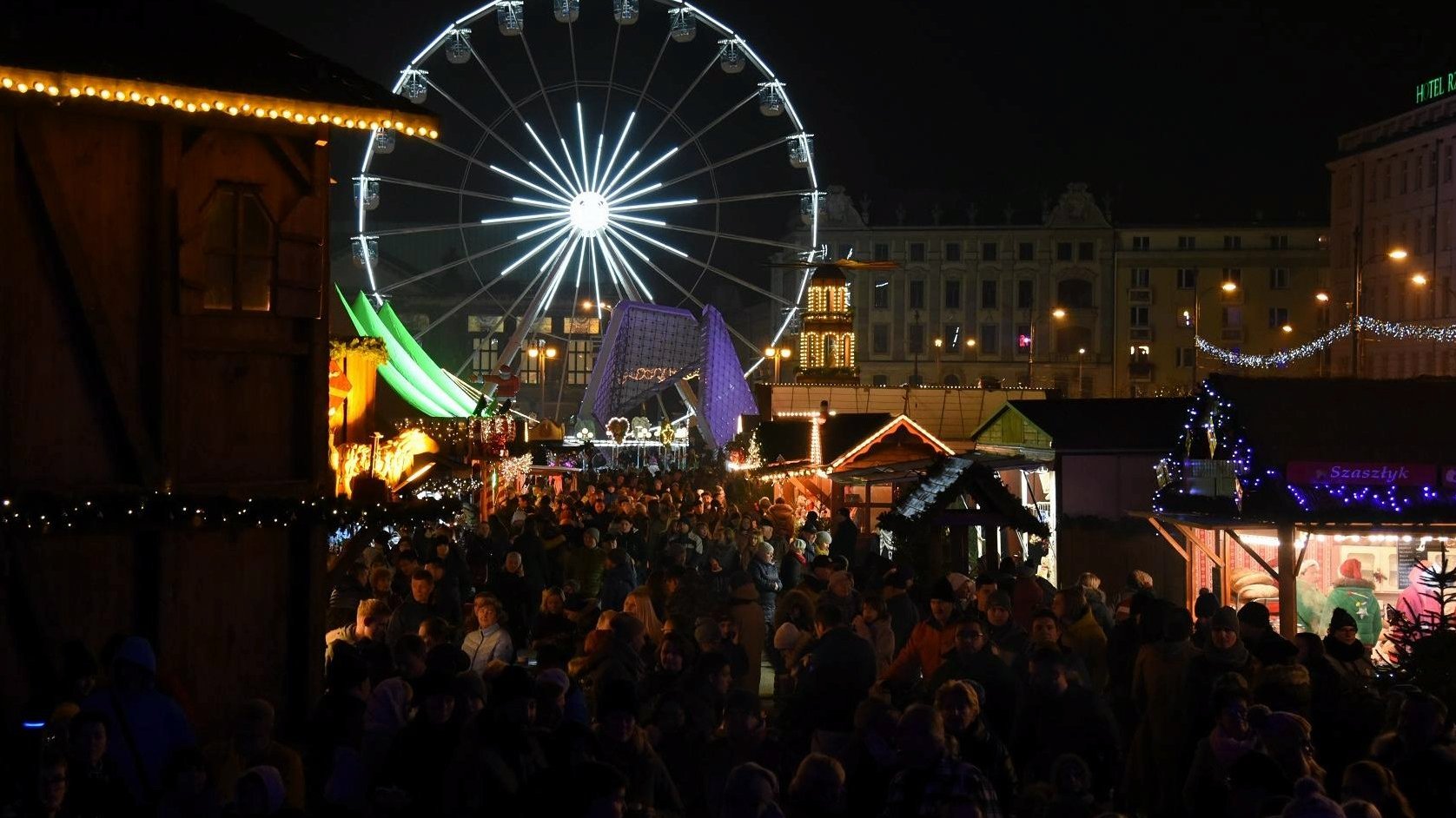 The photo shows crowd of people on the Christmas Fair, located on the Freedom Square, in evening scenery. In the background, behind several wooden stands, a ferris wheel with many bright, white lights is visible. - grafika artykułu