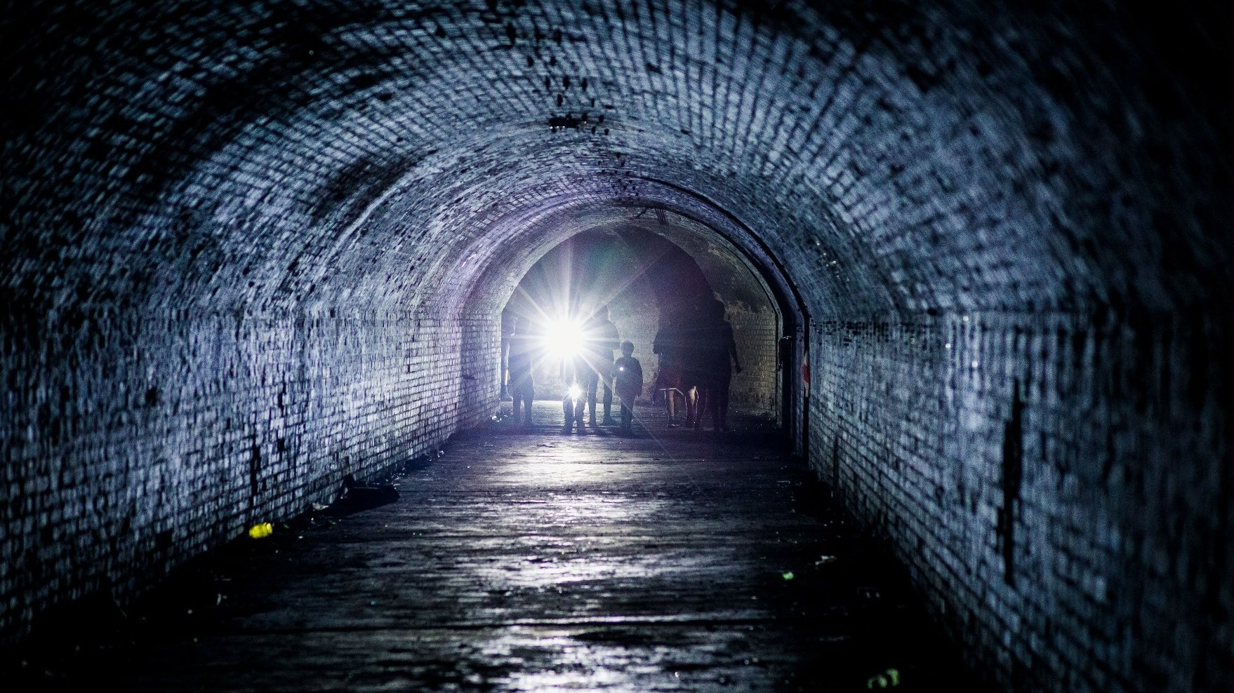 An underground corridor, at the end of which there are several people with flashlights. - grafika artykułu
