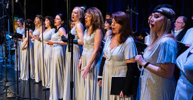 Photo from the concert: women in long white-silver dresses singing on stage.