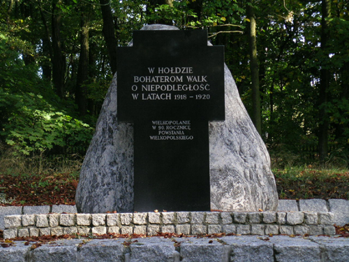 Duży kamień. Do kamienia przytwierdzona płyta w kształcie krzyża z napisem "W hołdzie Bohaterom walk o niepodległość w latach 1918-1920". W tle drzewa.