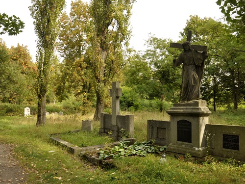 Najprawdopodobniej stare nagrobki. Na jednym duża figura Chrystusa z krzyżem na ramionach. Dookoła trawa i drzewa.