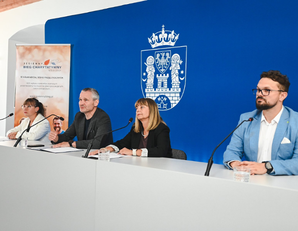 26 października na sportowej mapie Poznania pojawi się nowe wydarzenie - Jesienny Bieg Charytatywny Cztery osoby siedzą przy stole konferencyjnym podczas oficjalnej konferencji prasowej. Za nimi znajduje się niebieska ścianka z herbem miasta Poznania oraz flaga Unii Europejskiej. Po lewej stronie stoi roll-up promujący Jesienny Bieg Charytatywny. Osoby mają przed sobą mikrofony i szklanki z wodą, wyglądają na skupione i zaangażowane w omawiane tematy