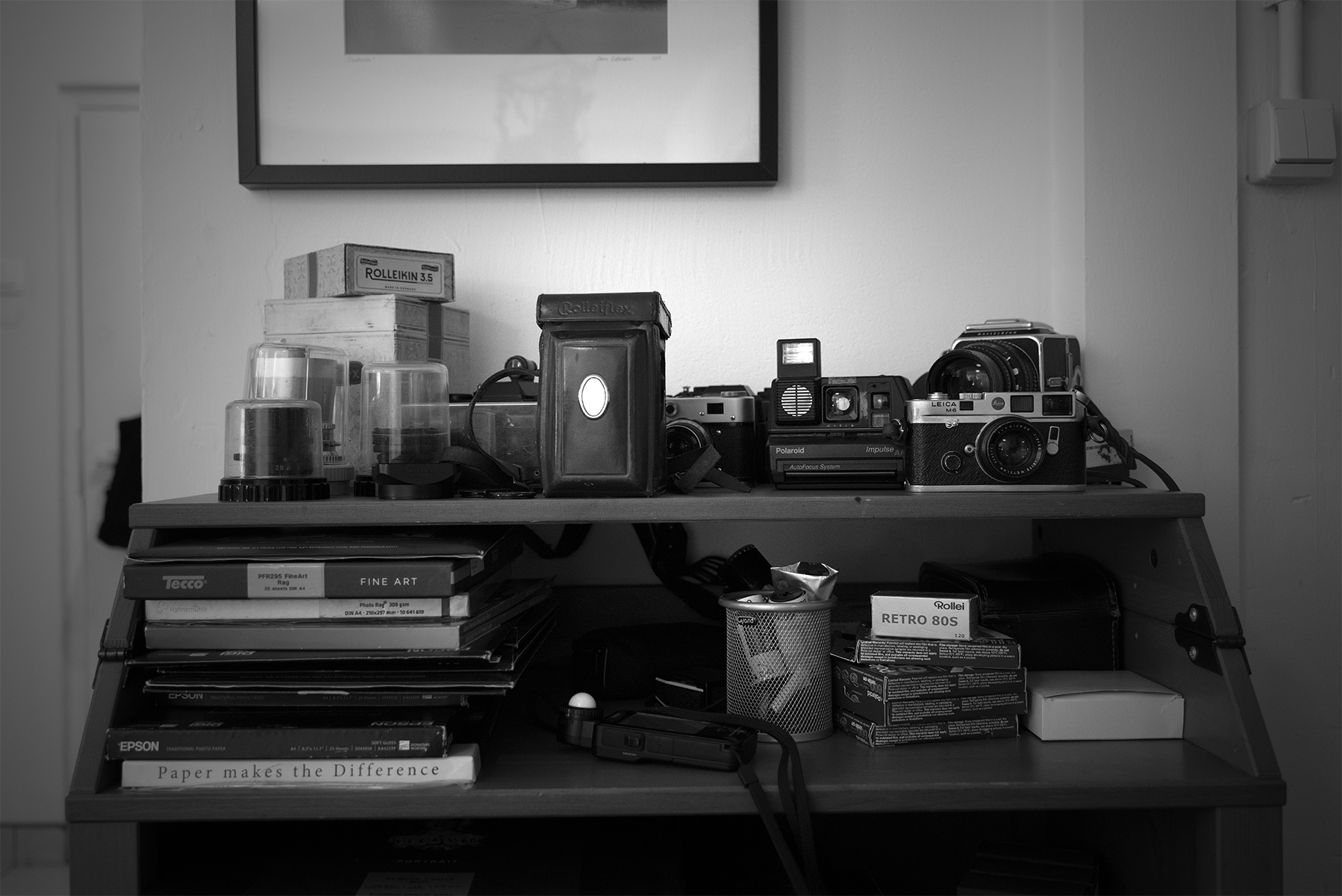 Zdjęcie przedstawia półki z albumami fotograficznymi oraz wyposażeniem pracowni: aparatami, światłomierzem, negatywami