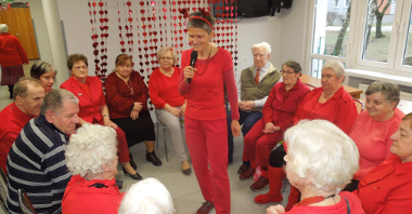 seniorzy ubrani na czerwono siedzą w kręgu na krzesłach i podają sobie czerwone serduszko