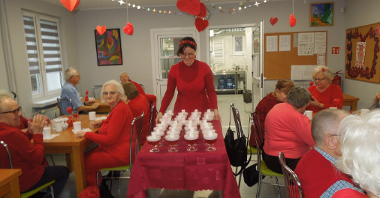 grupa seniorów siedzi przy stolikach, na środku sali stół na kółkach prowadzony przez kobietę ubraną na czerwono, nakryty bordowym obrusem, zastawiony szklanymi pucharkami z deserami,