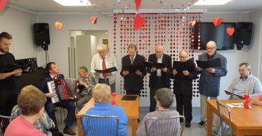 grupa panów (seniorów) stoi ze śpiewnikami w rękach, po lewej siedzi młody mężczyzna i gra na akordeonie