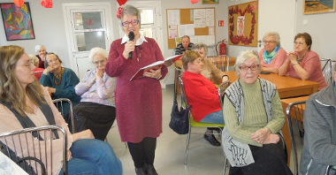 seniorka stoi na środku sali z mikrofonem w ręku, wokół siedzą przy stolikach starsze osoby