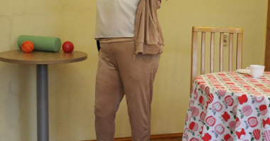 Zdjęcie przedstawia seniorkę w stroju sportowym, ćwiczącą z gumą.