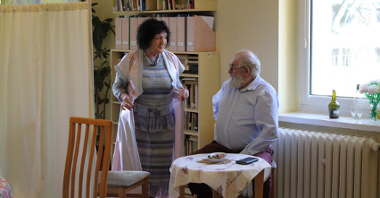 Na zdjęciu widać siedzącego przy stoliku mężczyznę oraz stojącą przy nim kobietę w szlafroku