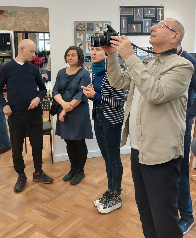 Otwarcie wystawy. Na pierwszym planie opiekun grupy fotograficznej z aparatem w rękach. Na dalszym planie opiekunka Galerii Starówka i kierowniczka DOA - grafika artykułu