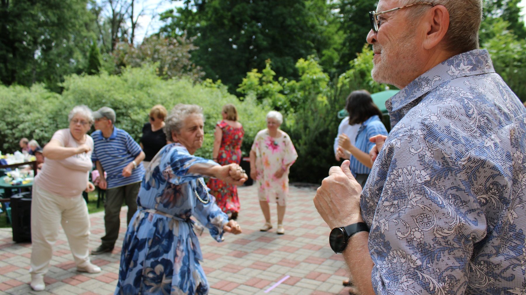 Na pierwszym planie uśmiechnięty mężczyzna w okularach wykonujący taneczne ruchy, po lewej seniorka w błękitnej zwiewnej sukni w duże granatowe kwiaty wygina się w rytm muzyki, w oddali więcej tańczących postaci w otoczeniu zieleni