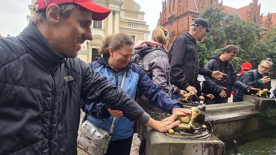 Na zdjęciu widać grupę zwiedzających Toruń stojących dookoła fontanny , którą okalają złote żaby. Kilka osób głaszcze metalowe żaby zgodnie z wierzeniem ,ze przyniesie to spełnienie ich marzeń. W tle budynki starówki Torunia.