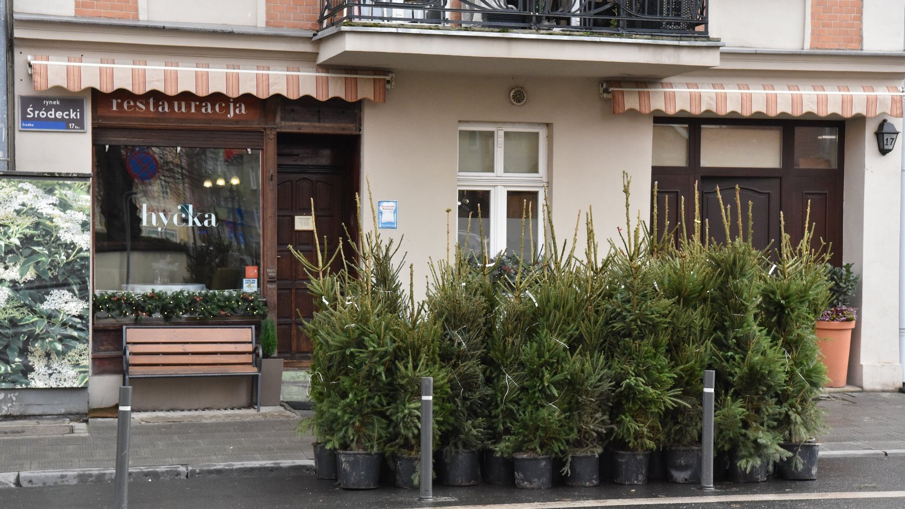 Zewnętrzna część restauracji o nazwie 'Hyćka'. Restauracja ma markizę w paski i szyld z napisem 'restauracja' nad wejściem. Przed restauracją, na chodniku, ustawiono kilka doniczek z drzewkami iglastymi. Przed oknem restauracji znajduje się również ławka. Po lewej stronie restauracji znajduje się znak uliczny z napisem 'rynek Śródecki'. Budynek ma balkon na górnym piętrze z czarną poręczą