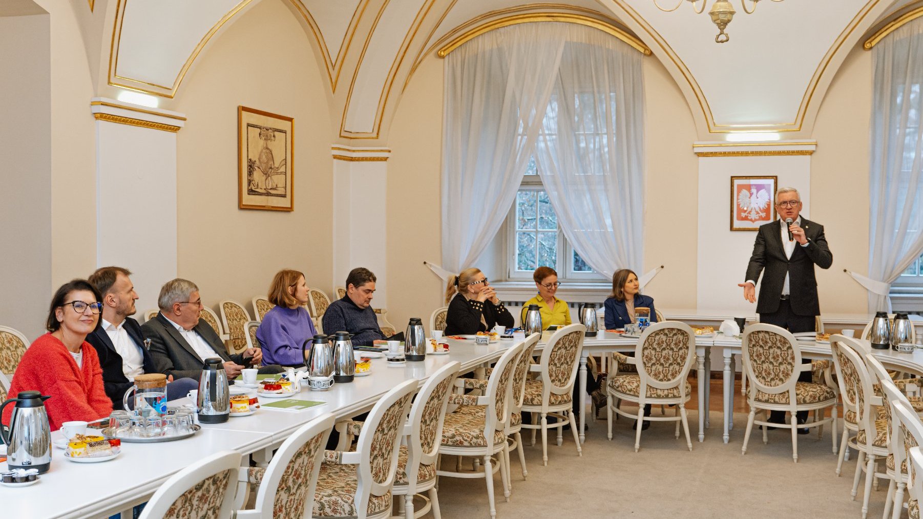 sala z ozdobnymi, złotymi łukami na ścianach i krzesłami. Przy długim stole siedzą kobiety i mężczyźni (Radni Miasta Poznania), przed nimi termosy z kawą oraz poczęstunek. Na szczycie stołu stoi prezydent Jaśkowiak ubrany w garnitur. Trzyma w ręku mikrofon i mówi coś do niego.