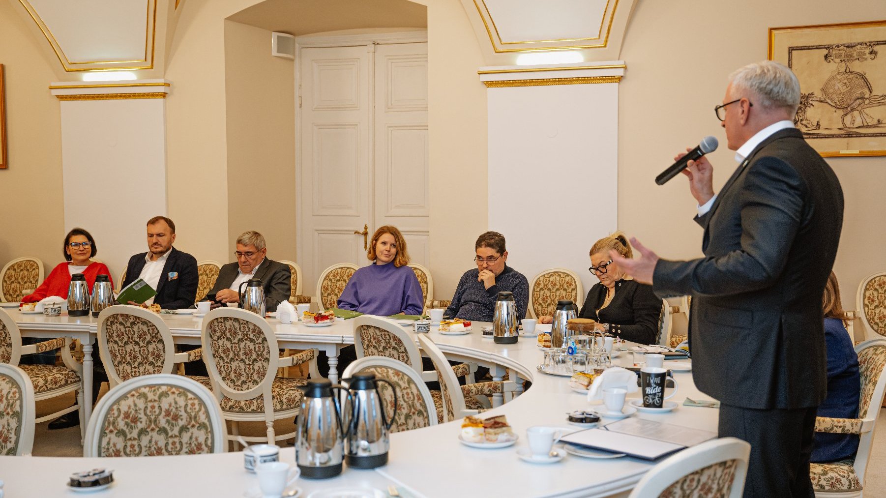 sala z ozdobnymi, złotymi łukami na ścianach i krzesłami. Przy długim stole siedzą kobiety i mężczyźni (Radni Miasta Poznania), przed nimi termosy z kawą oraz poczęstunek. Na szczycie stołu stoi prezydent Jaśkowiak ubrany w garnitur. Trzyma w ręku mikrofon i mówi coś do niego.