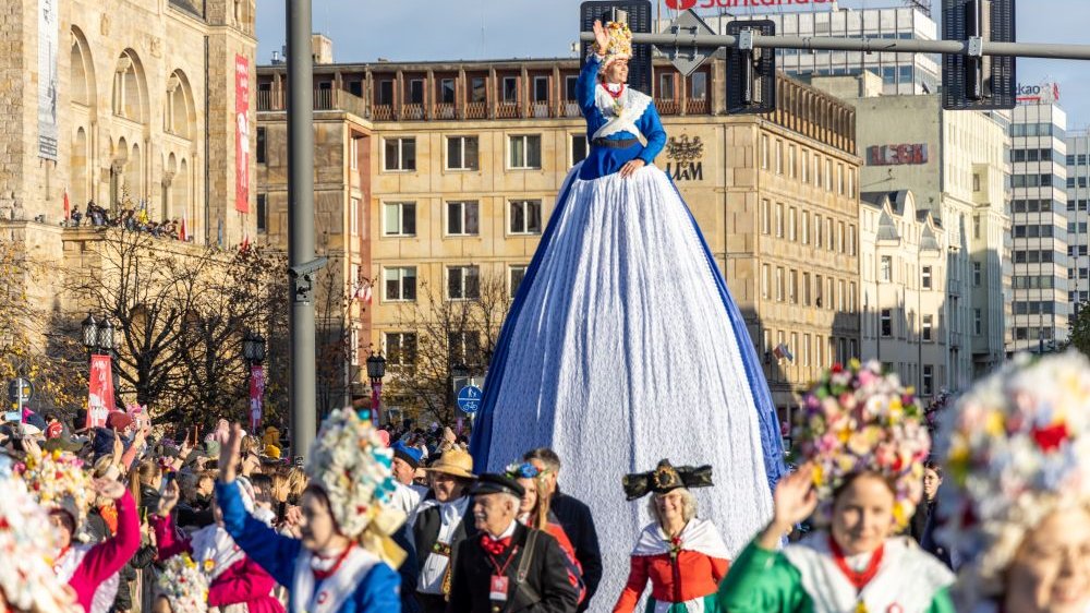 Kobieta w stroju bamberki na wysokiej platformie przejeżdża ulicą w pochodzie. Macha ręką do ludzi. Za jej plecami budynek CK Zamek i dawnego Collegium Historicum.