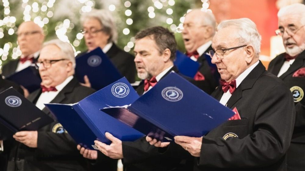 Elegancko ubrani mężczyźni z czerwonymi muchami śpiewają w chórze. W tle widoczna ozdobiona światełkami choinka.