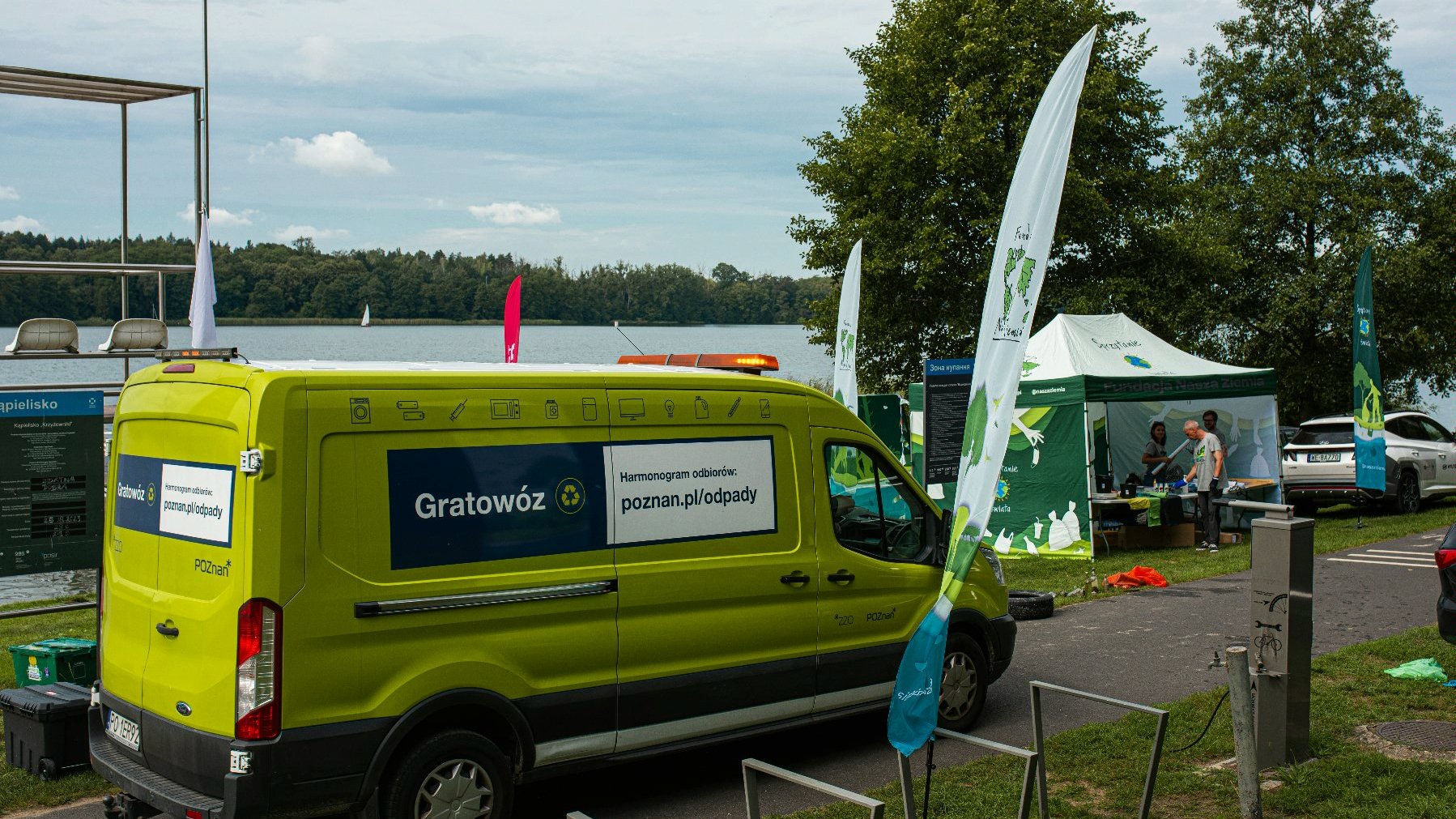 Na fotografii widać zielony, duży pojazd - Gratowóz, przed nim jest miejsce na pozostawienie rowerów, w tle widać namiot Fundacji Nasza Ziemia opisany tematycznie - Sprzątanie Świata, a za nim znajduje się jezioro. - grafika artykułu