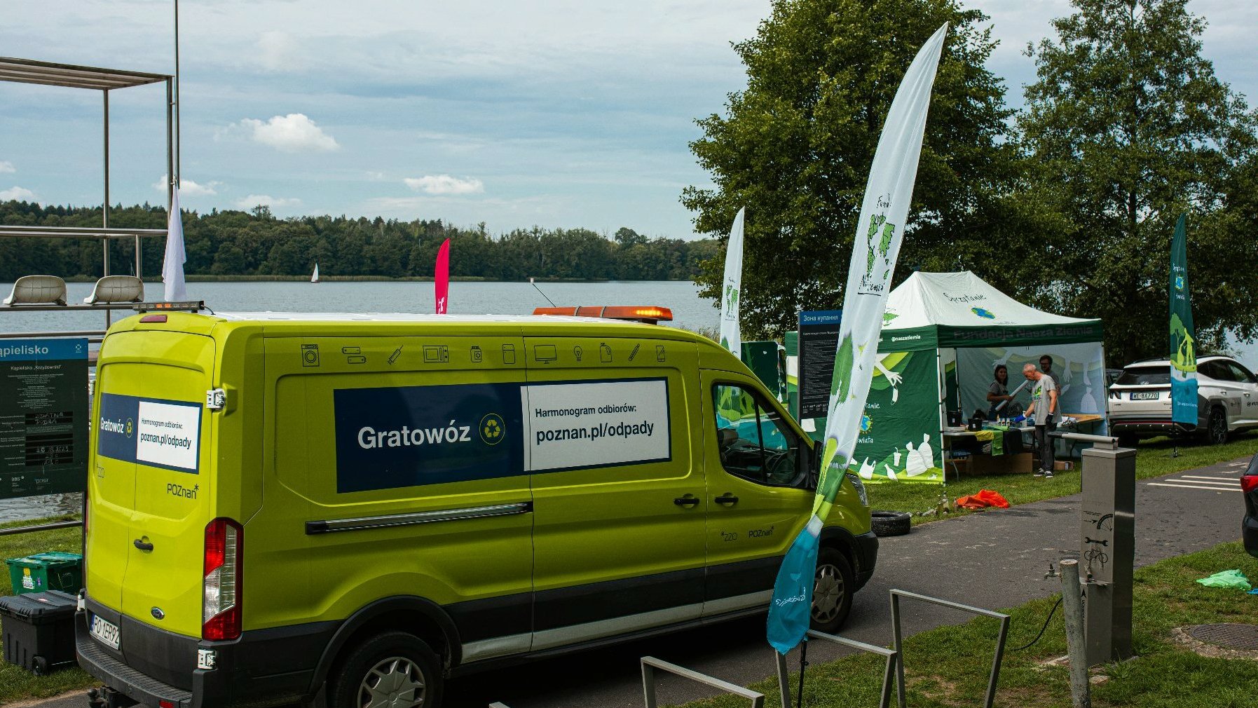 Na fotografii widać zielony, duży pojazd - Gratowóz, przed nim jest miejsce na pozostawienie rowerów, w tle widać namiot Fundacji Nasza Ziemia opisany tematycznie - Sprzątanie Świata, a za nim znajduje się jezioro.