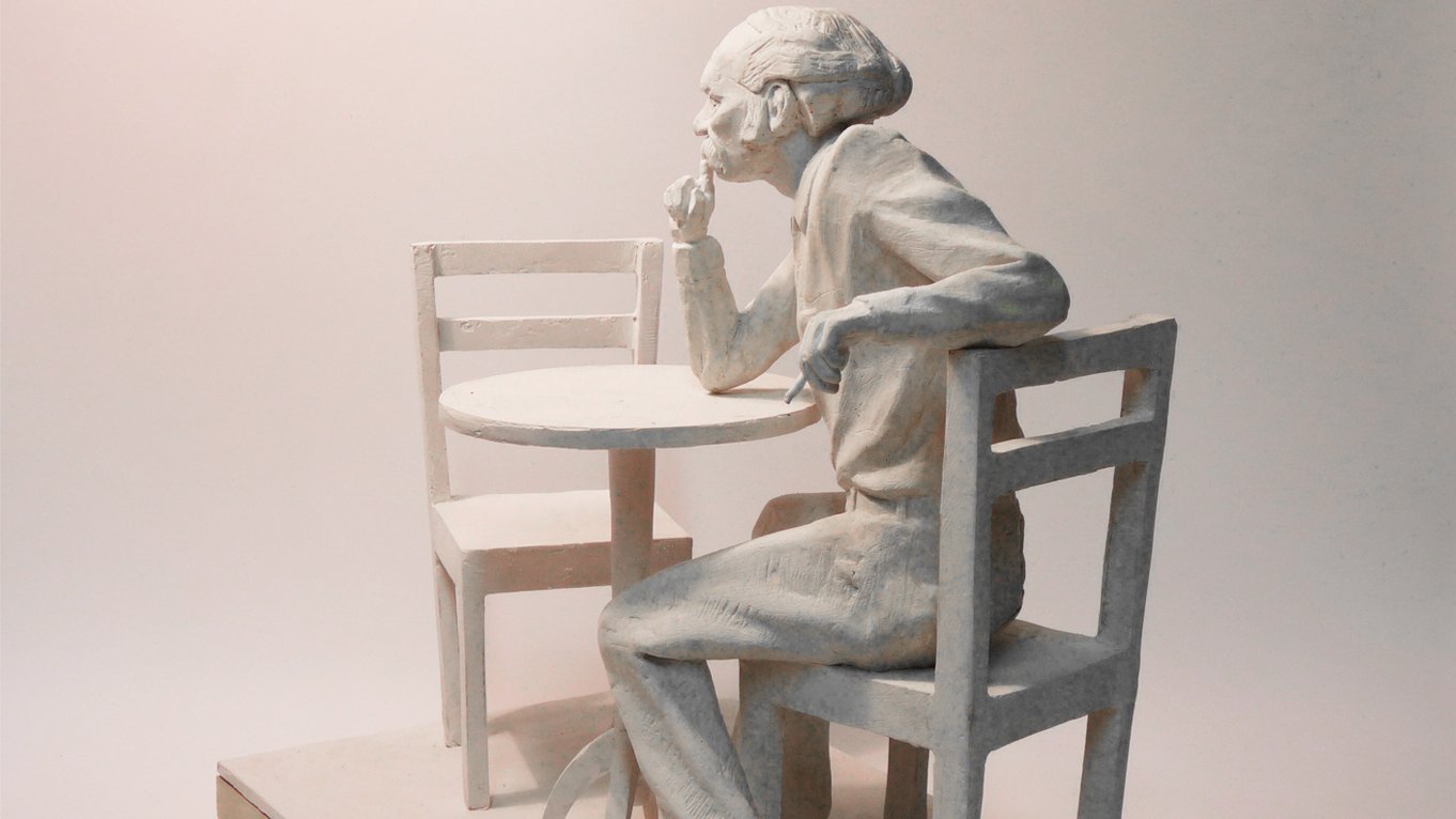 Projekt: Bohdan Smoleń siedzący na krześle przy niewielkim stoliku, zamyślony. Prawy łokieć opiera na stole i dłonią dotyka brody, lewa dłoń na oparciu krzesła. Naprzeciw drugie, puste krzesło