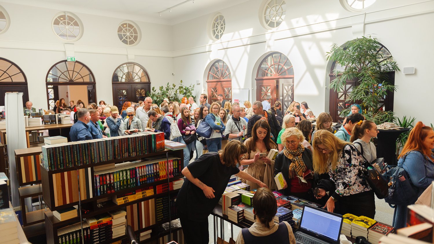 Tłum ludzi w sali, gdzie jest dużo książek. Stoją na półkach i leżą na ladzie. Ludzie oglądają i kupują książki.