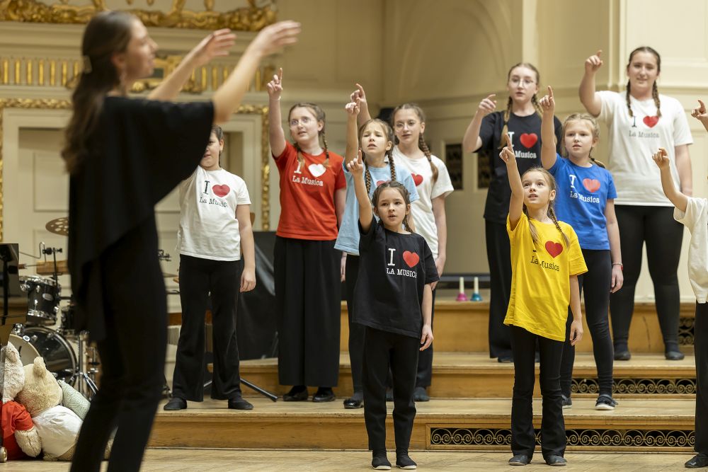 Na zdjęciu dzieci i młodzież na próbie chóru, ubrani w kolorowe t-shirty, podnoszą prawe ręce w górę. Naprzeciwko nich stoi dyrygentka. - grafika artykułu