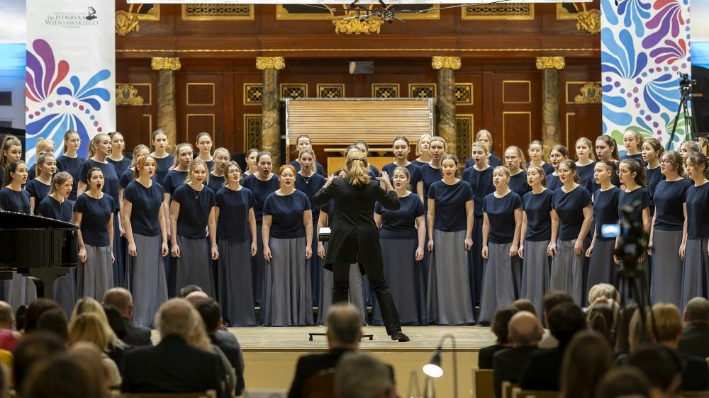 Na zdjęciu widoczny chór żeński na scenie podczas koncertu, naprzeciwko dyrygentka