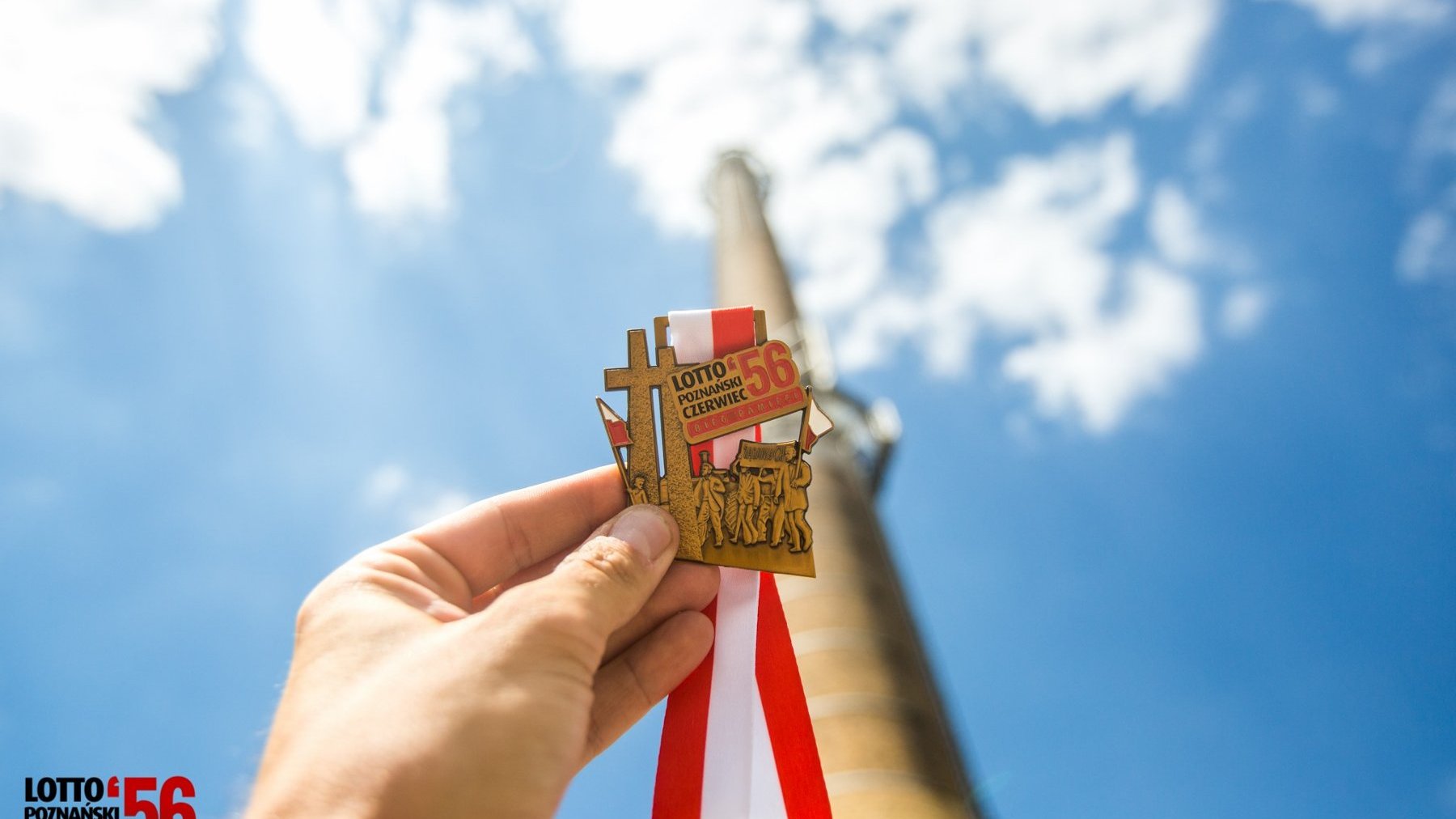 Dłoń trzyma medal z okazji Biegu Pamięci Poznańskiego Czerwca '56. W tle wysoki komin hutniczy, niebieskie niebo i chmury.