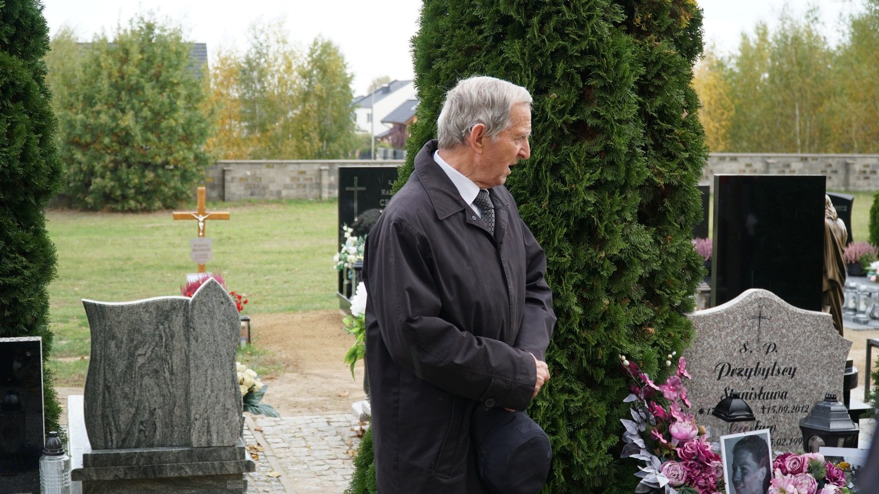 Starszy mężczyzna z siwymi, krótkimi włosami, ubrany z długi ciemny płaszcz. Spogląda na grób zmarłej Stanisławy Sobańskiej i wygłasza przemowę.