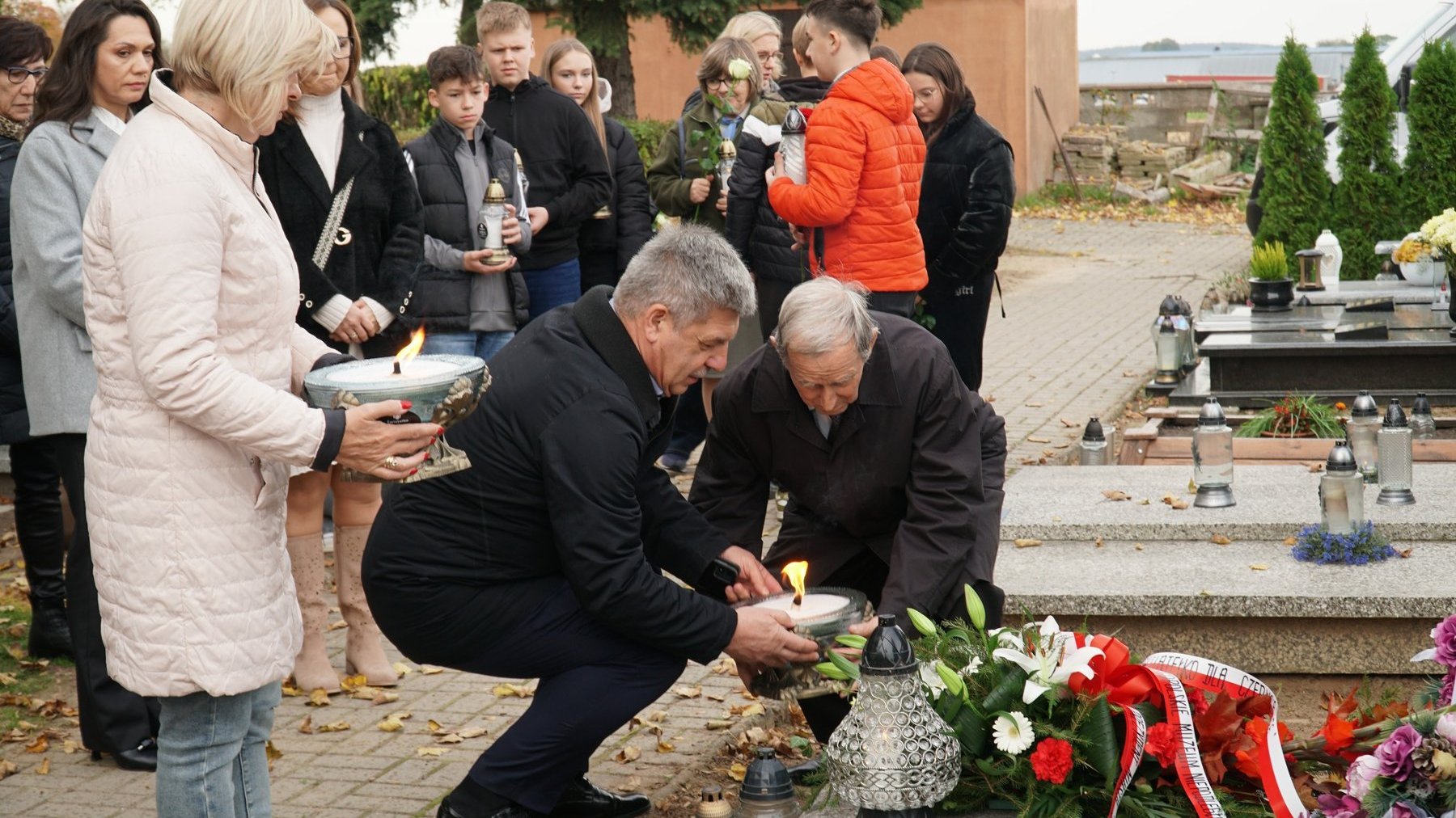 Dwóch mężczyzn w podeszłym wieku pochyla się nad grobem i zapala symboliczny znicz. Obok stoi kobieta w średnim wieku i trzyma drugi znicz. W tle pozostali uczestnicy przyglądają się.